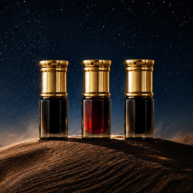 Three perfume bottles with gold caps stand on a desert sand dune under a starry night sky. The bottles contain dark and red liquids, with dramatic lighting highlighting their metallic caps and glass bodies.